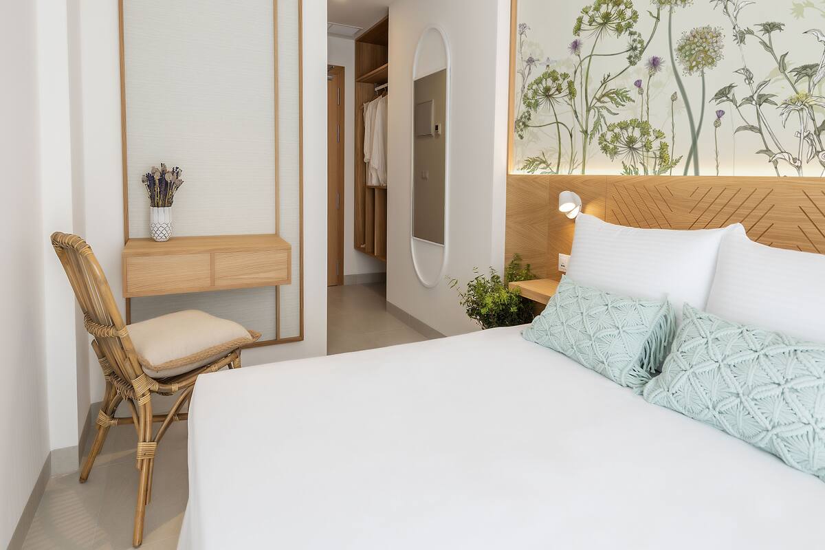 Une chambre à coucher comprenant un lit, une chaise, une table de chevet, un miroir et un mur orné d’un motif floral.