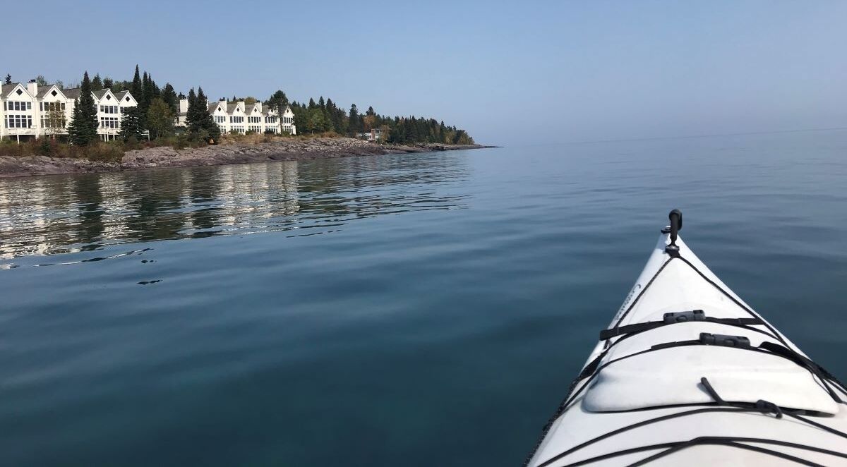 Un kayak sobre aguas tranquilas, con una orilla que presenta edificios blancos y árboles.
