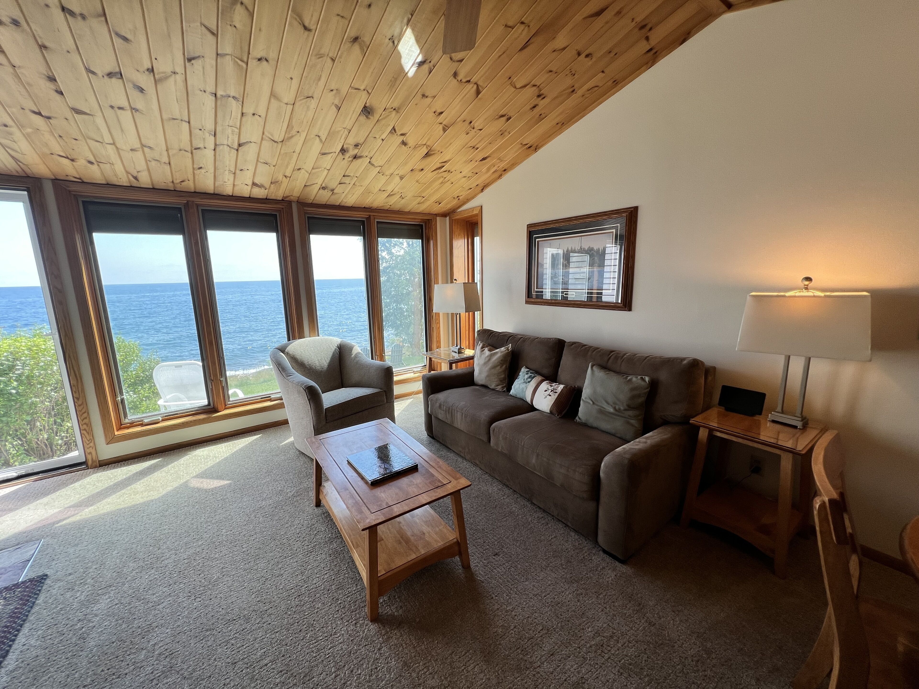 Una sala de estar acogedora con techo de madera, ventanas amplias con vistas al mar, un sofá, un sillón y una mesita.