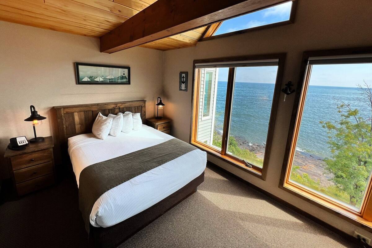 A bedroom with a large bed, wooden headboard, bedside lamps, a nightstand, and a view of the ocean through large windows.