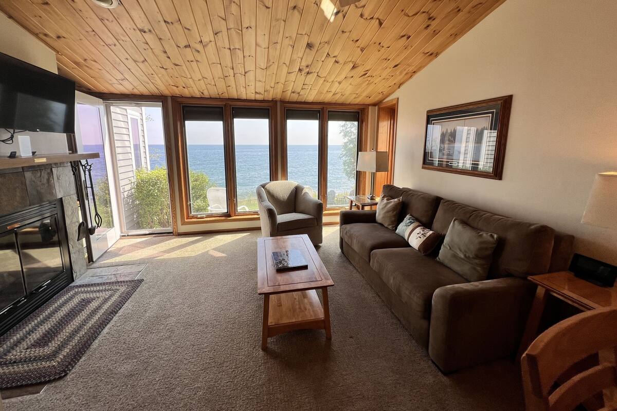 A cozy living room with a wooden ceiling, a fireplace, a sofa, a coffee table, and a large window offering a view of the sea.