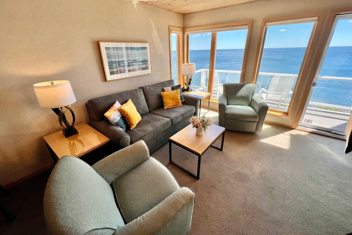 A modern living room with a sofa, armchairs, a coffee table, and a view of the ocean through large windows.