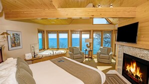 Une chambre spacieuse avec un grand lit, une cheminée, une télévision et une vue sur la mer grâce à de grandes fenêtres.