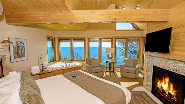 Un dormitorio amplio con una cama grande, chimenea, televisor y vista al mar a través de amplias ventanas.