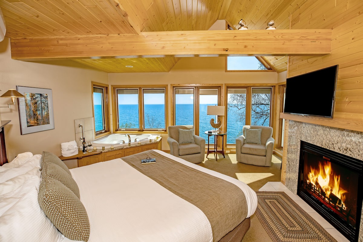 A spacious bedroom with a large bed, a fireplace, a TV, and a view of the sea through large windows.