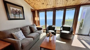 Sala de estar con techo de madera, un sofá gris, dos sillones, una mesa de centro con un jarrón y ventanas amplias con vista al mar.
