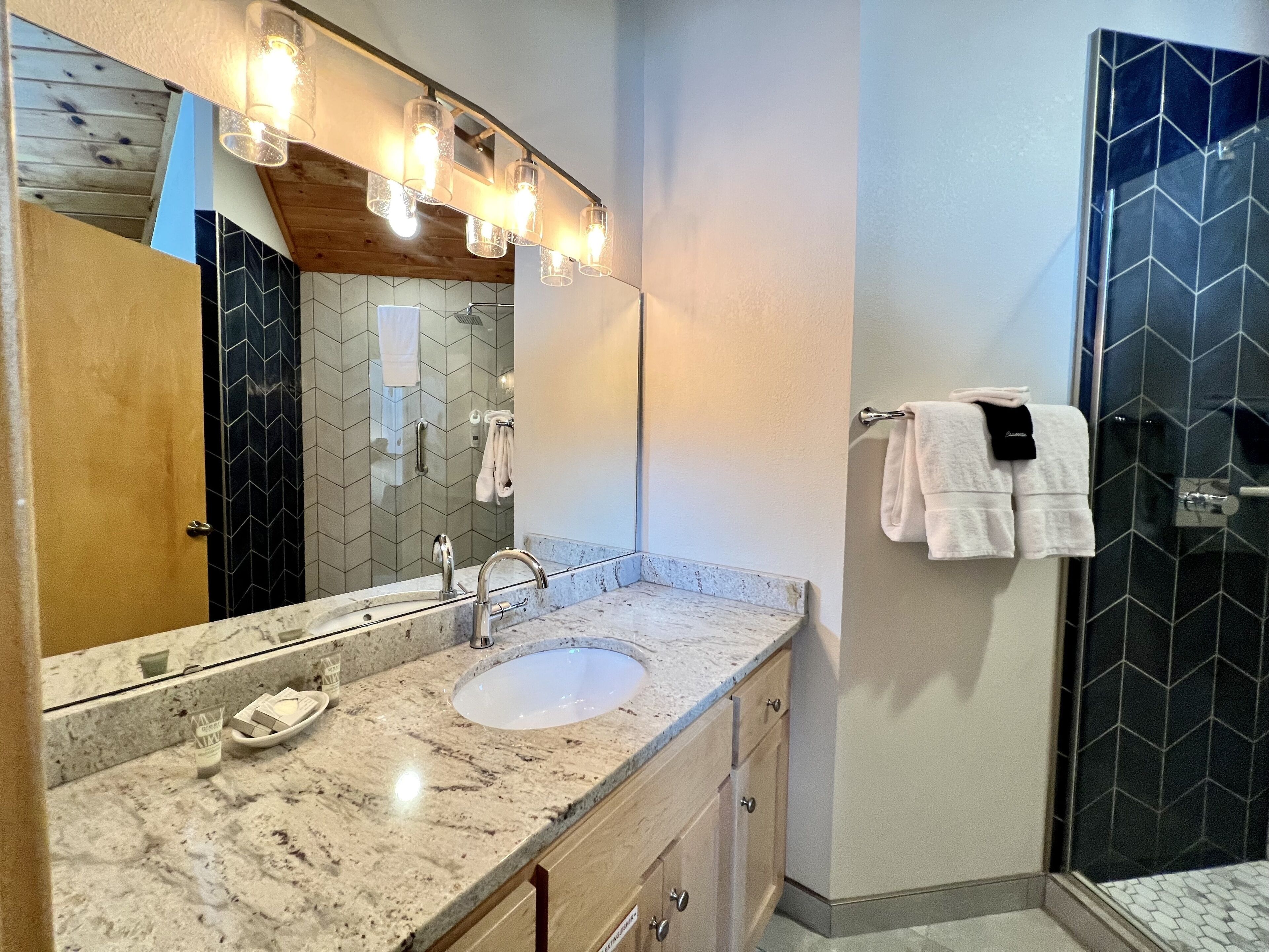 Un baño con lavamanos de mármol, un espejo grande y una ducha con azulejos negros estampados.
