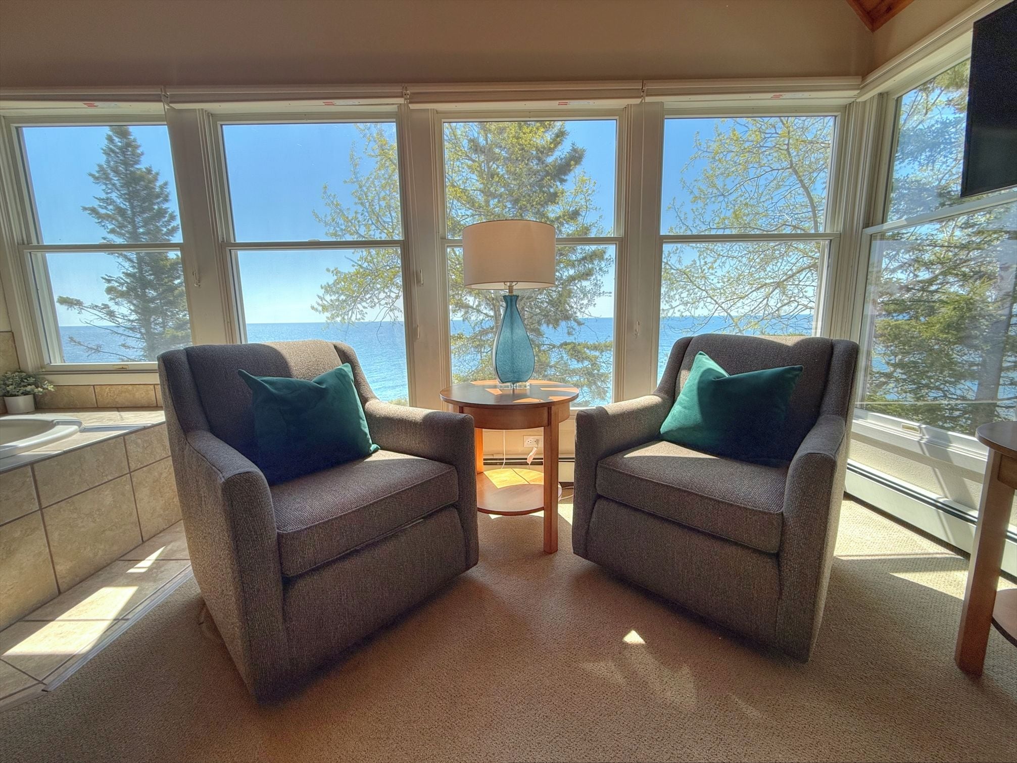 A sunroom with two armchairs, a small table with a lamp, and a view of the sea through large windows.