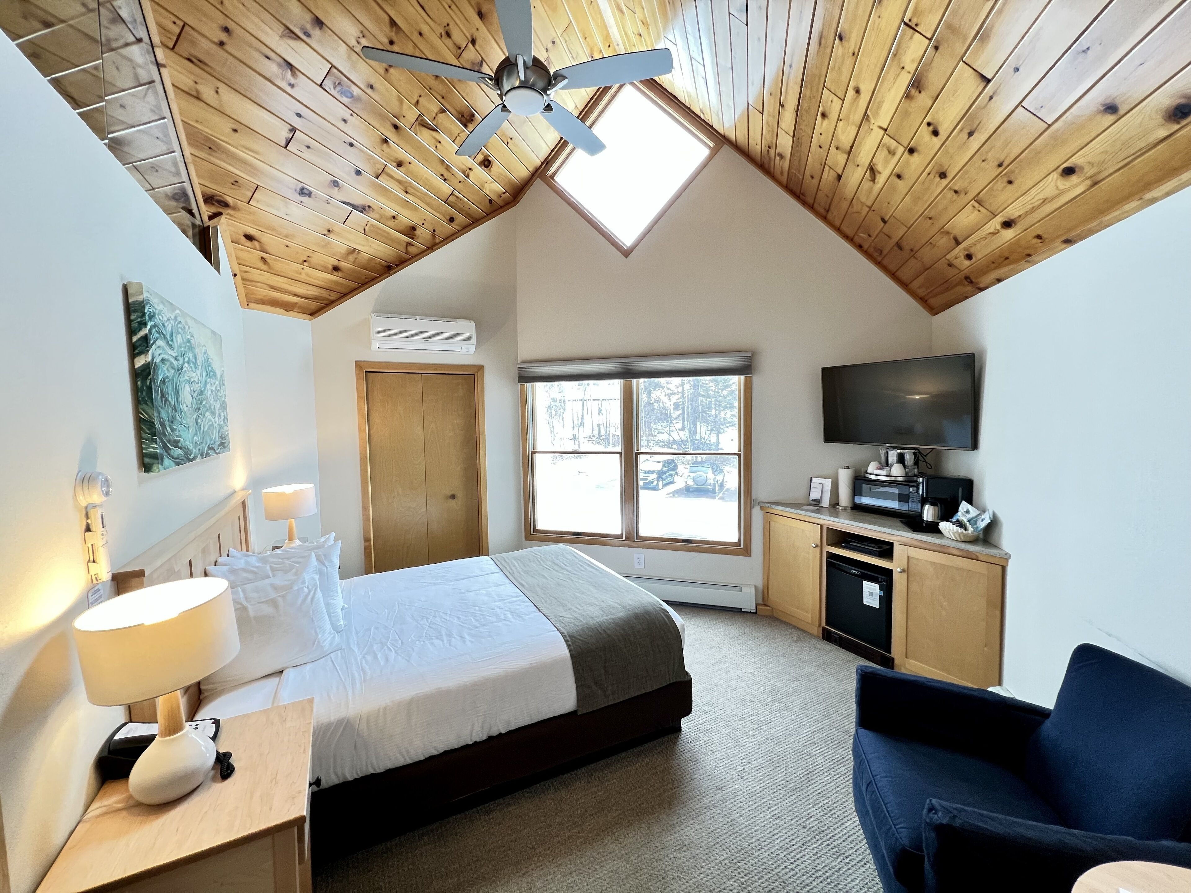 Une chambre à coucher comprenant un lit, un bureau, une chaise, un téléviseur et une fenêtre donnant sur des arbres et une voiture.