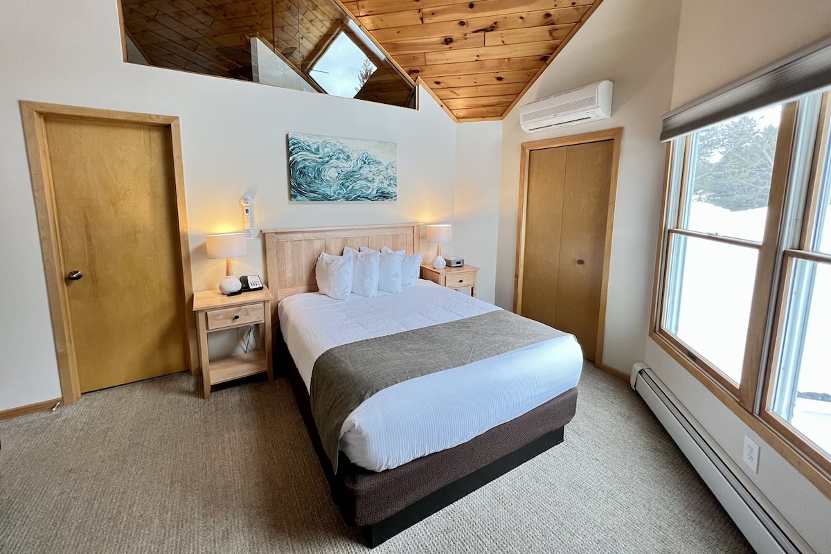 A bedroom with a wooden ceiling, a large bed, bedside lamps, a painting, and a window.