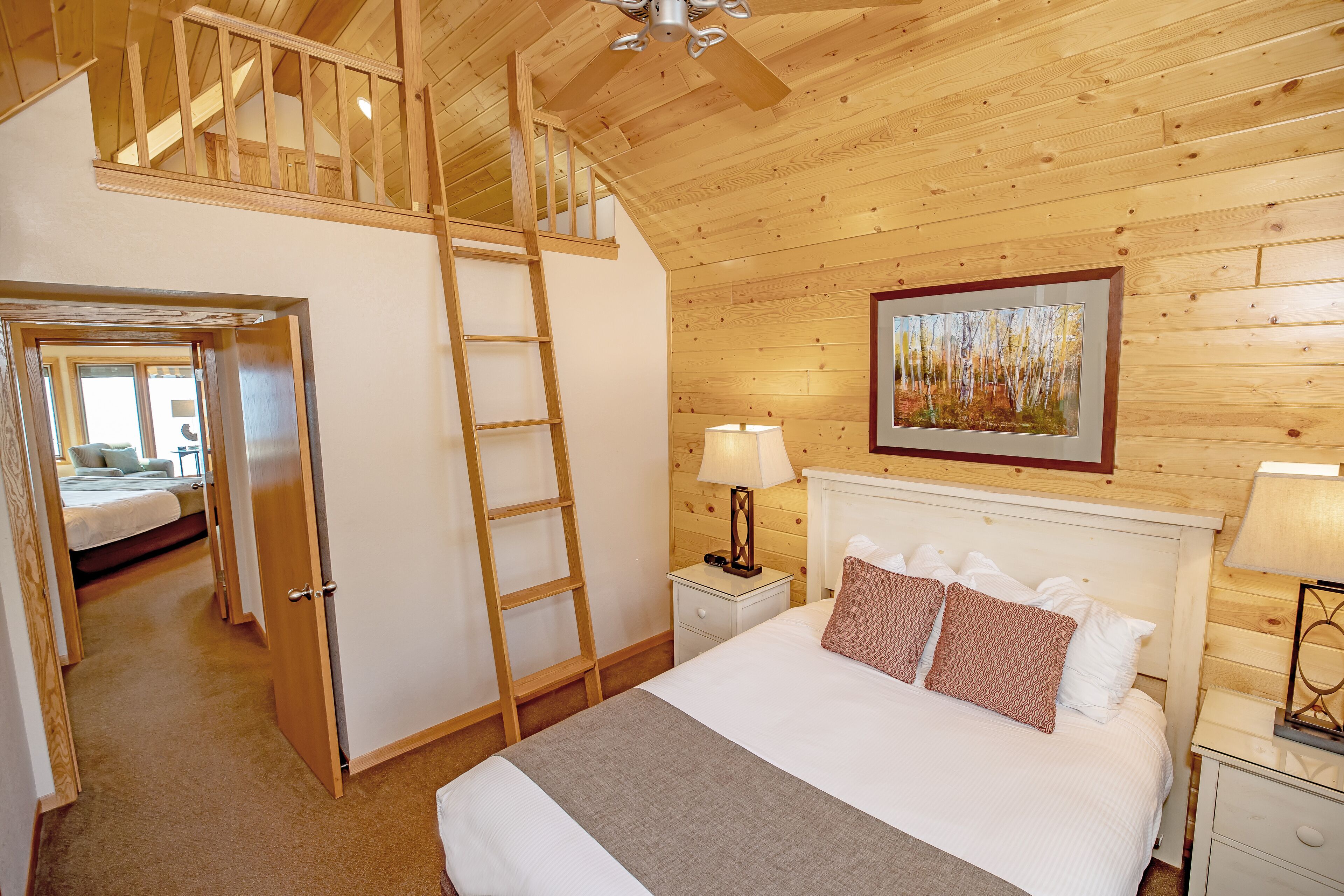 Un dormitorio con techo estilo loft, una escalera de madera, una cama con almohadas blancas y estampadas, una mesita de noche con lámparas y un cuadro en la pared.