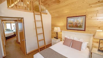 Un dormitorio con techo estilo loft, una escalera de madera, una cama con almohadas blancas y estampadas, una mesita de noche con lámparas y un cuadro en la pared.