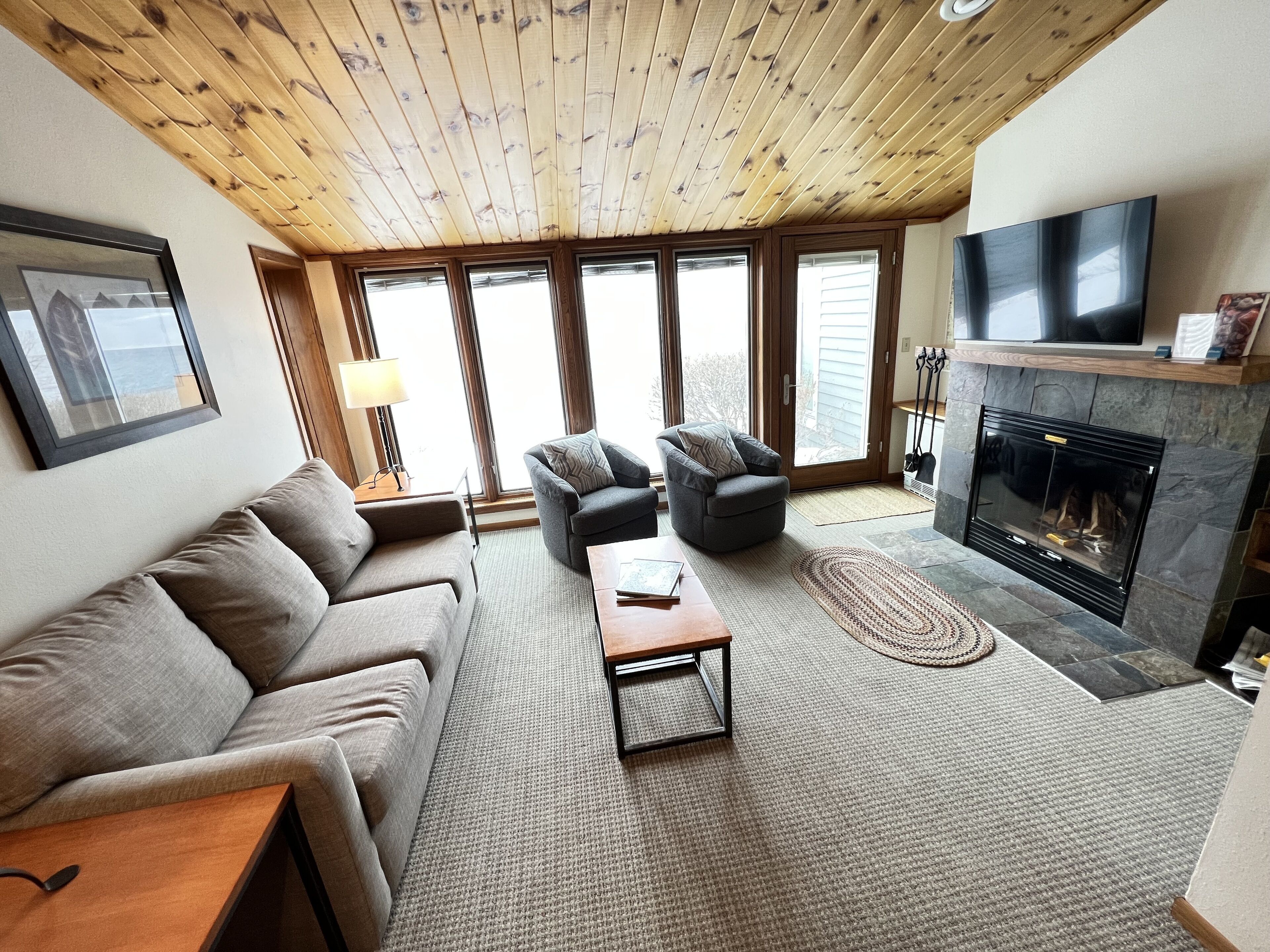 Una sala de estar acogedora con techo de madera, chimenea, sofá, sillón y televisor.