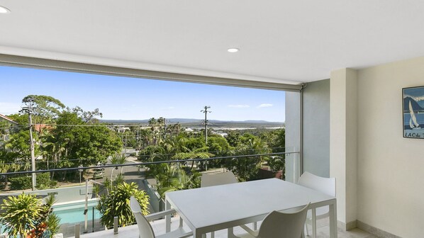 A balcony with a pool, a table, and chairs, overlooking a lush landscape and distant hills.