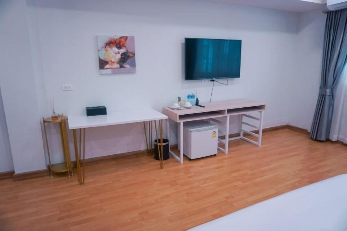 A room with a white desk, a flat-screen TV, a small refrigerator, and a painting on the wall.