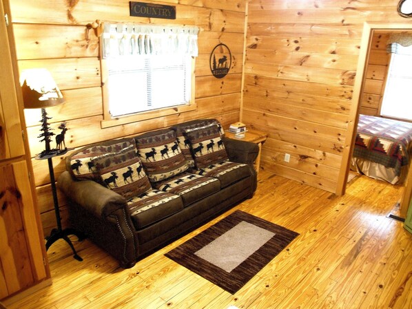 A wooden cabin interior with a sofa, a lamp, and a window with blinds. - Moose Cabin - 2 Bedroom/1 Bath Vacation Rental (Franklin)