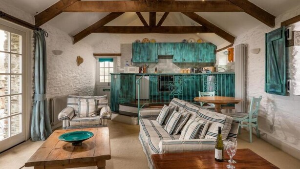 A rustic room with exposed wooden beams, a striped sofa, a wooden table, and a kitchen with teal cabinets. - Tregarland Farm (Looe)