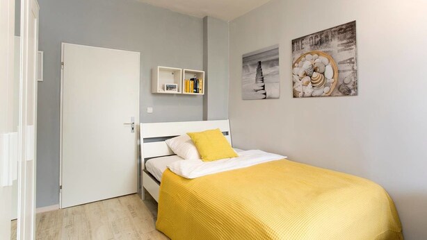 Un dormitorio con cama, una puerta blanca, una estantería con libros y dos cuadros en la pared.