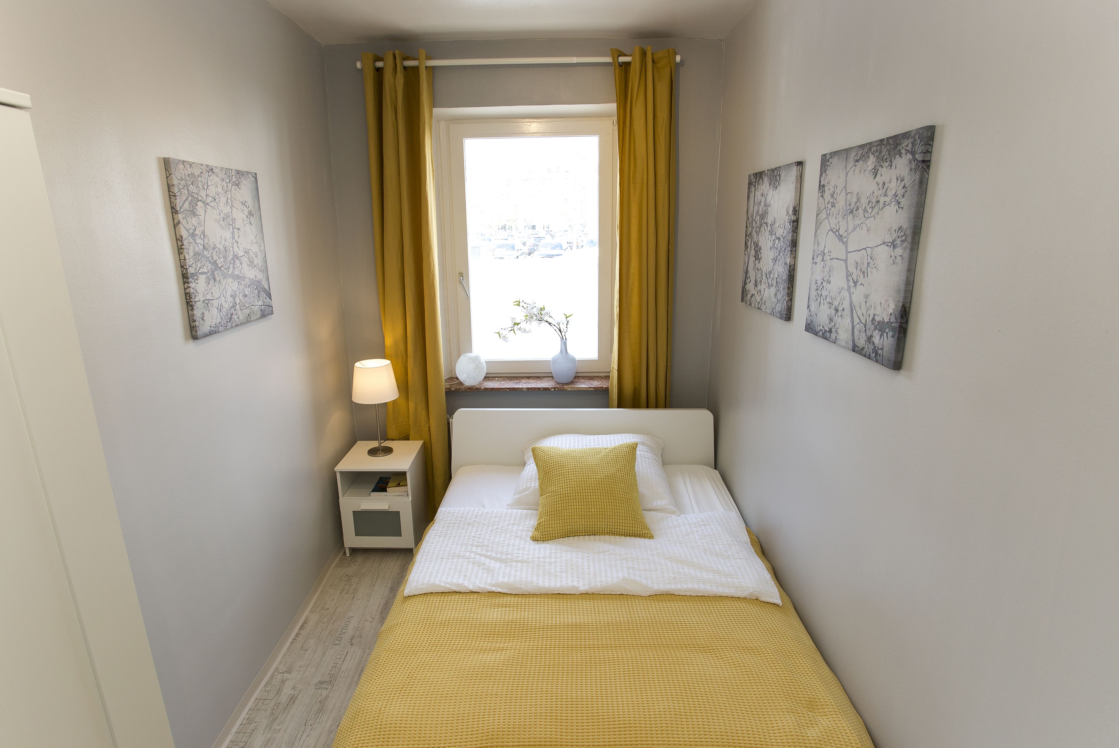 A bedroom with a bed, a nightstand, a lamp, a window with curtains, and two wall-mounted artworks.
