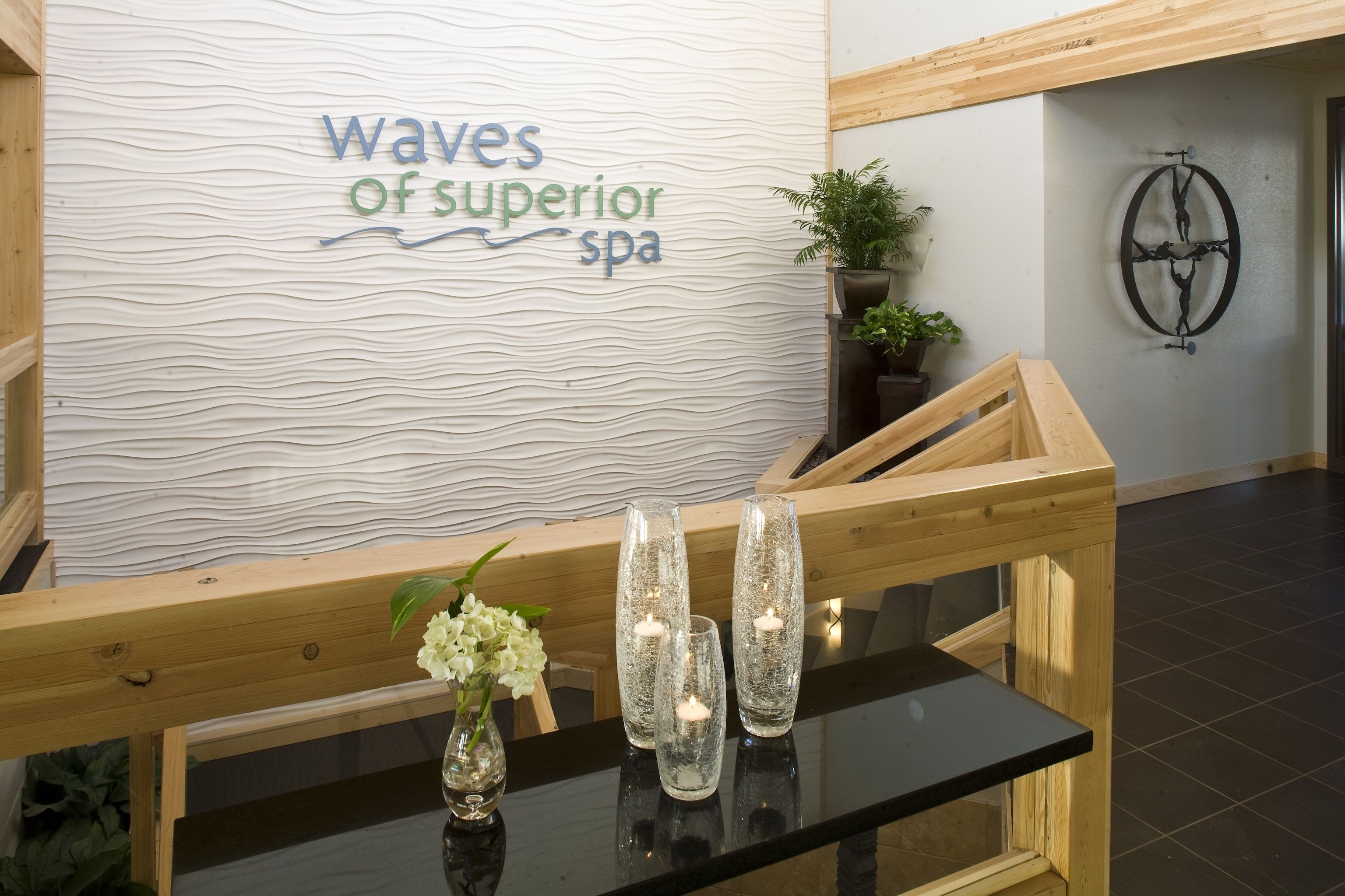 Aula, jossa on puinen penkki, lasipöytä maljakkoineen ja kynttilöineen, seinä, jossa on kyltti, jossa lukee ”Waves of superior spa”, ruukkukasvi ja seinäkello.