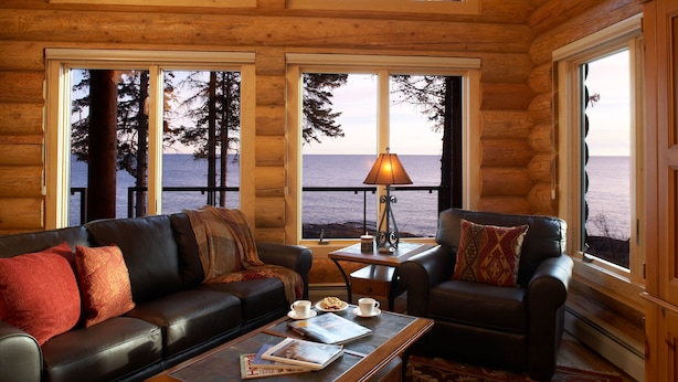 A log cabin interior with a leather sofa, a coffee table, and a view of the water through large windows. - Temperance Landing on Lake Superior (Schroeder)