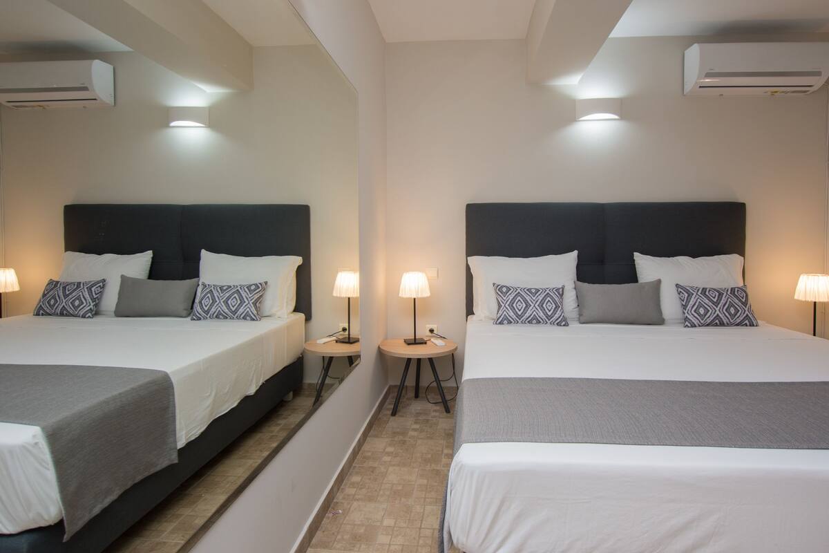 A hotel room with two beds, each with a black headboard, white bedding, and patterned pillows. There are two bedside tables with lamps, and the room has a tiled floor.