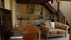 Ein gemütliches Wohnzimmer mit einer Couch und einem Esstisch mit Stühlen. Im Hintergrund ist eine Küche mit einer Mikrowelle und Schränken zu sehen.