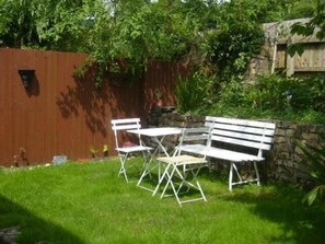 A small outdoor seating area with a white metal table and bench set against a stone wall, surrounded by lush greenery.