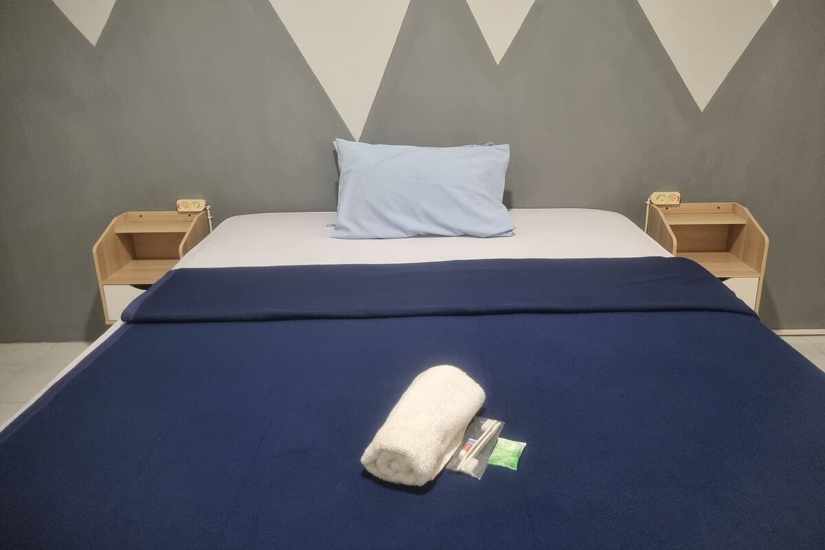A neatly made bed with a blue bedspread, a white pillow, and a rolled towel on the bed. There are two wooden nightstands with a small lamp and a power outlet on each.