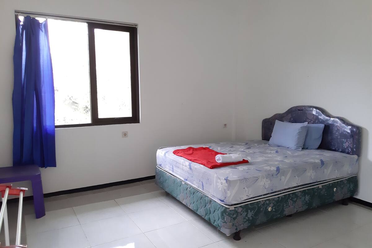 A bedroom with a bed, a window with blue curtains, and a small table with a chair.