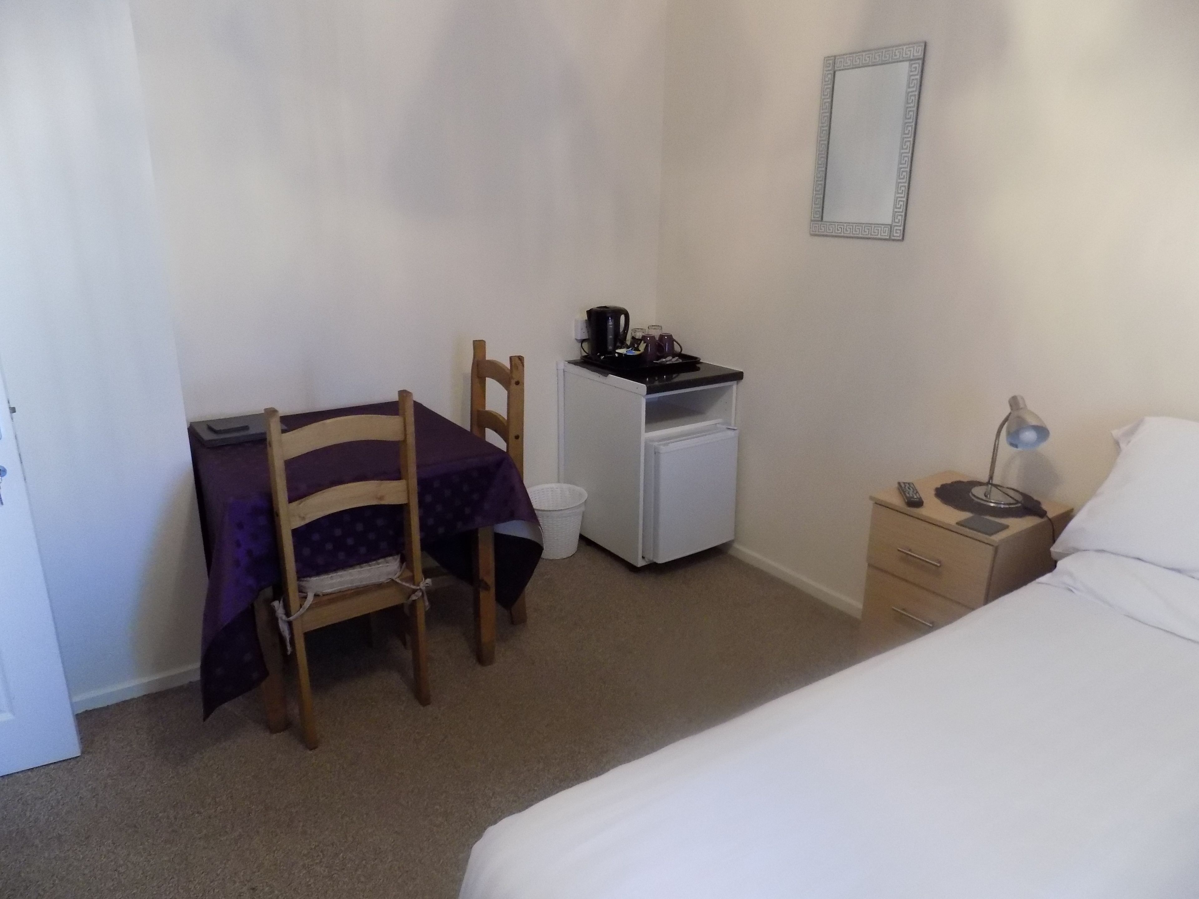 Een compacte, netjes ingerichte slaapkamer met een bed, een bureau met stoel, een kleine koelkast, een nachtkastje met lamp en een spiegel aan de muur.