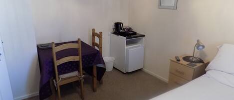 A small, neatly arranged bedroom with a bed, a desk with a chair, a small refrigerator, a nightstand with a lamp, and a wall-mounted mirror.