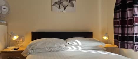 A hotel room with a bed, two bedside lamps, a wall-mounted fan, and a painting.