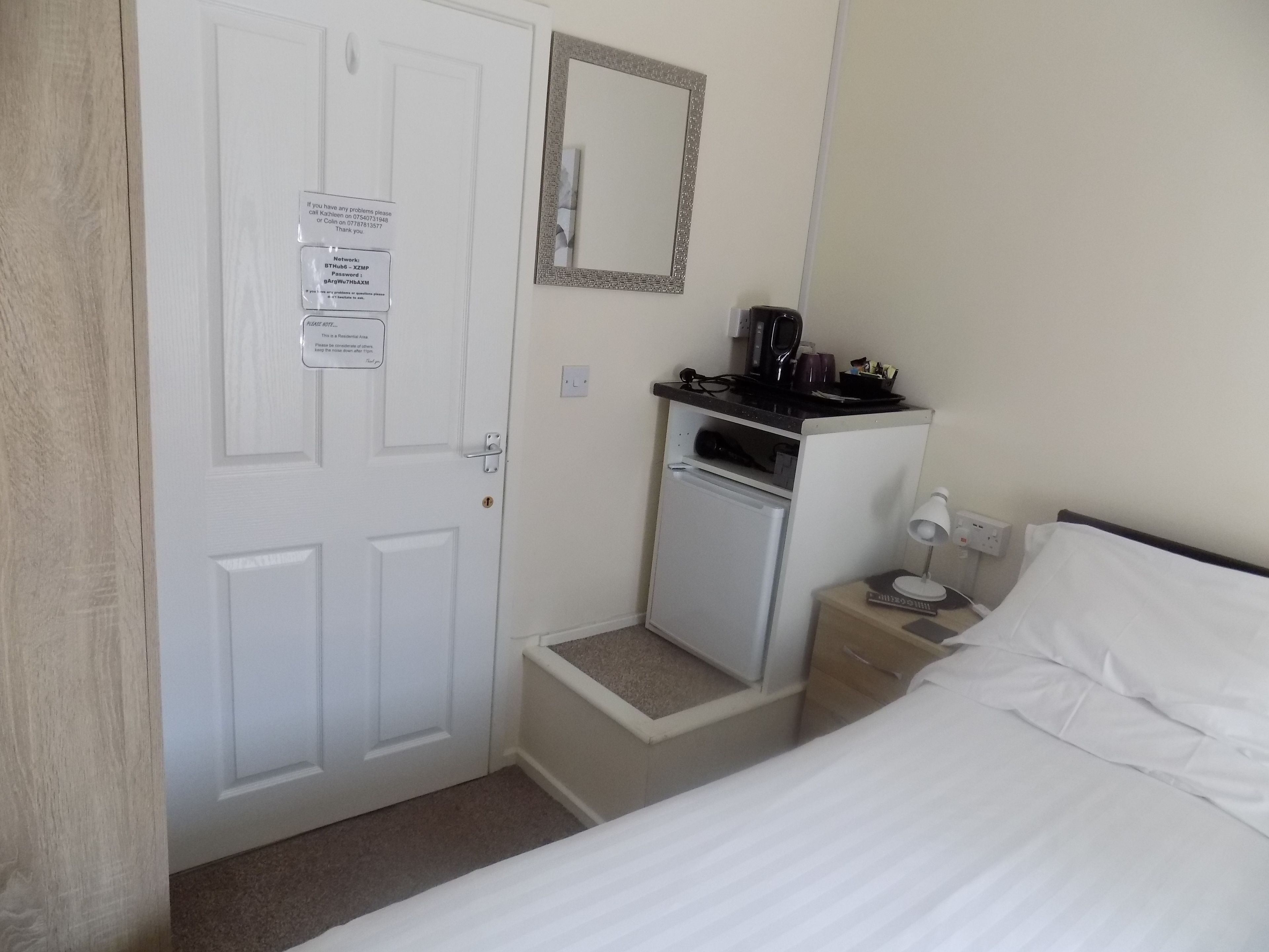 A small, neatly organized room with a bed, a nightstand, a mirror, and a door with a notice.
