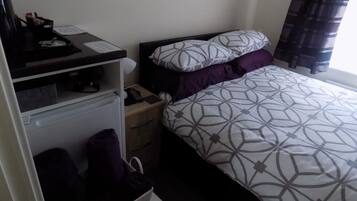 A bedroom with a bed, a nightstand, a dresser, and a desk with a computer and a lamp.