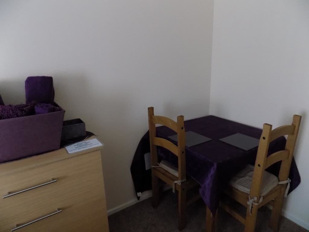 Une petite pièce avec un meuble en bois, deux chaises en bois et une table recouverte d’une nappe violette.