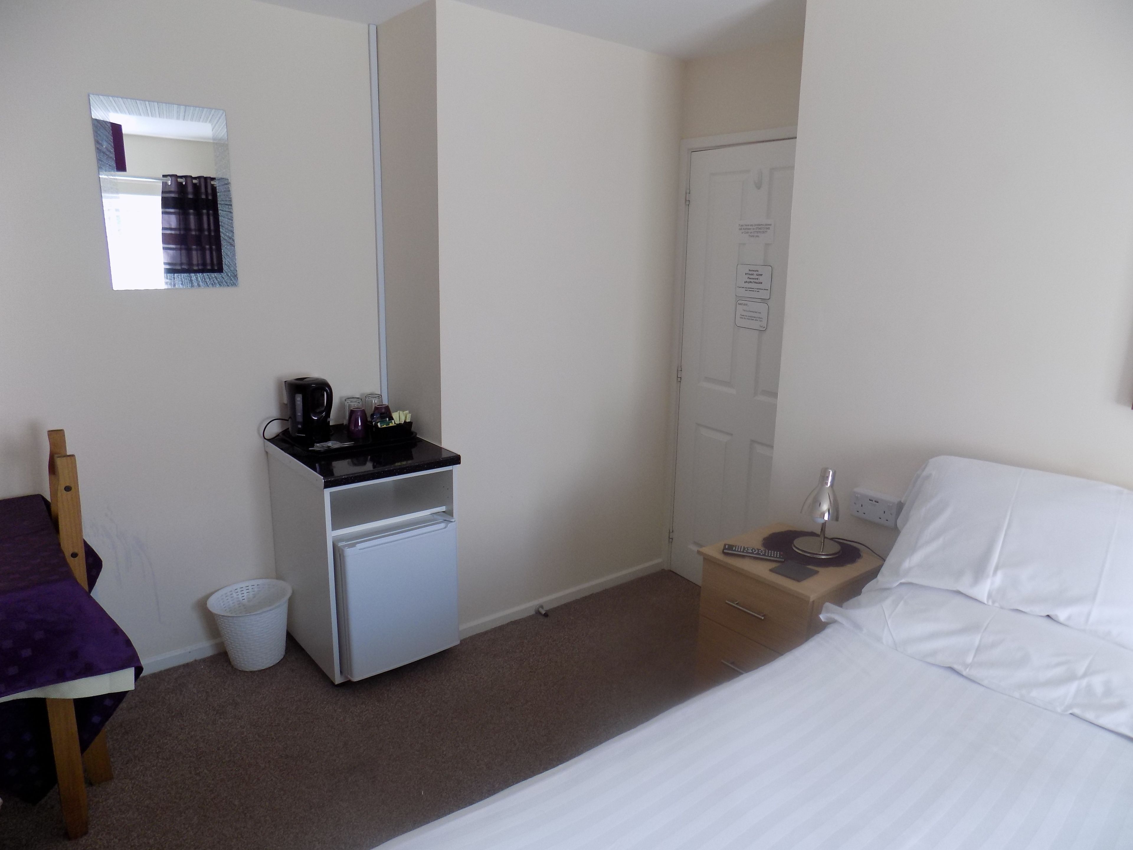 A hotel room with a bed, a desk with a coffee maker, a trash bin, and a door with a sign.