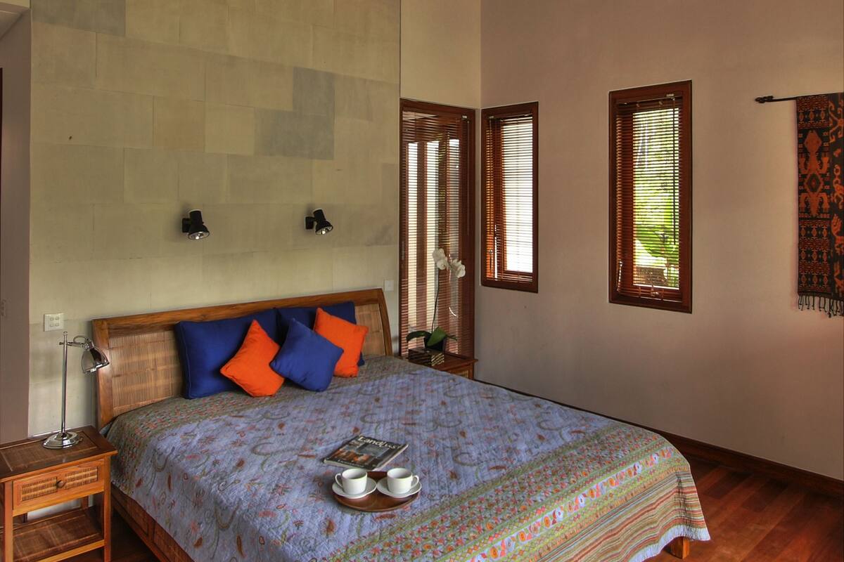 Une chambre avec un plafond en bois, un grand lit avec une literie à motifs, deux lampes fixées au mur et une fenêtre avec des stores.