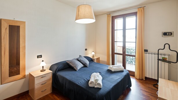 A bedroom with a bed, bedside lamps, a wooden nightstand, a mirror, a window with curtains, and a radiator. - Lucca - Via Viaccia 155 (Lucca)