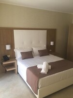 Una cama bien hecha con un cabecero marrón, dos lámparas de noche, un teléfono y una toalla enrollada sobre la cama.