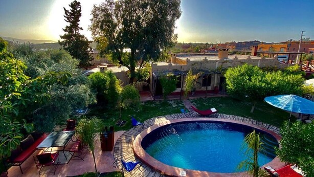 A swimming pool with a wooden deck, surrounded by greenery and a view of buildings in the background. - Relais Esmeralda (Lalla Takarkoust)