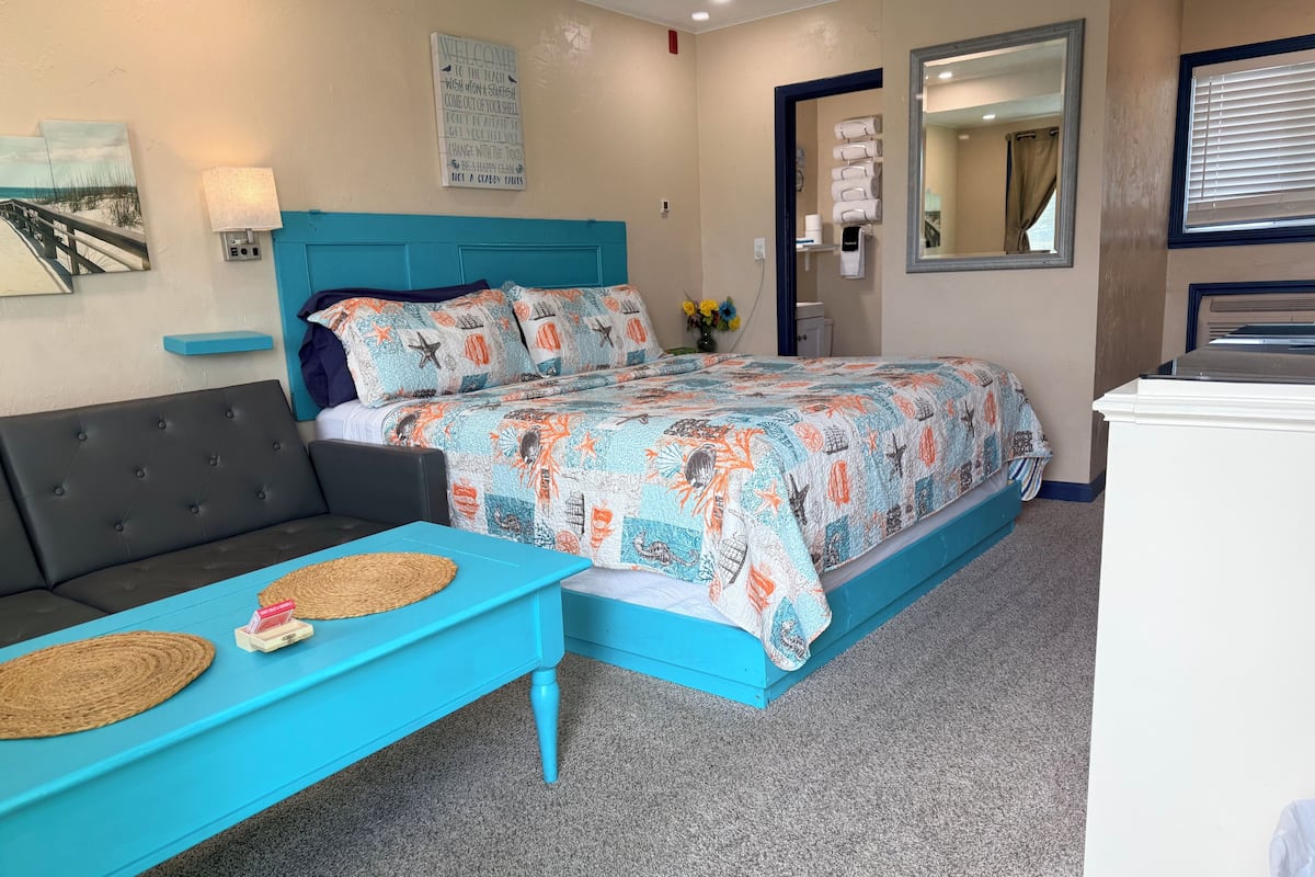Um quarto com cama, um sofá azul, uma mesinha, uma luminária na parede, um espelho e uma placa de boas-vindas.