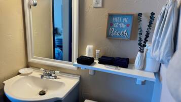 A bathroom with a white sink, a mirror, a towel rack with towels, a 'Meet Me at the Beach' sign, and a potted plant.