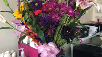 Un escritorio con un florero morado lleno de flores coloridas, un tarro de vidrio con caracoles, una botella de jabón de manos, un bloc de notas y una superficie de escritorio azul.