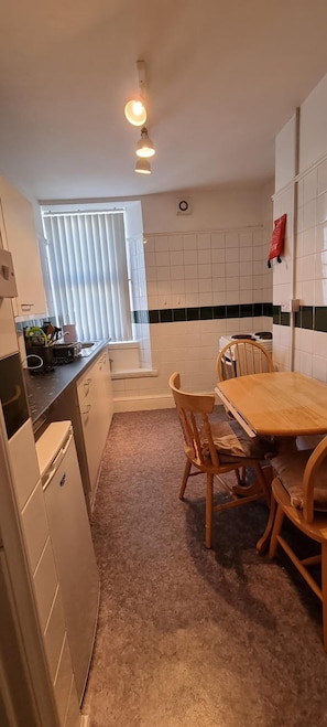 A compact kitchen with a dining table and chairs, a refrigerator, and a window with blinds. - Comfort Rooms (Plymouth)