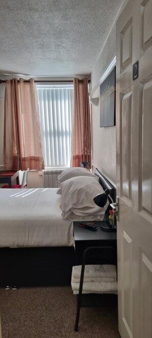 A hotel room with a bed, a desk with a lamp, a chair, a radiator, and a window with curtains. - Comfort Rooms (Plymouth)