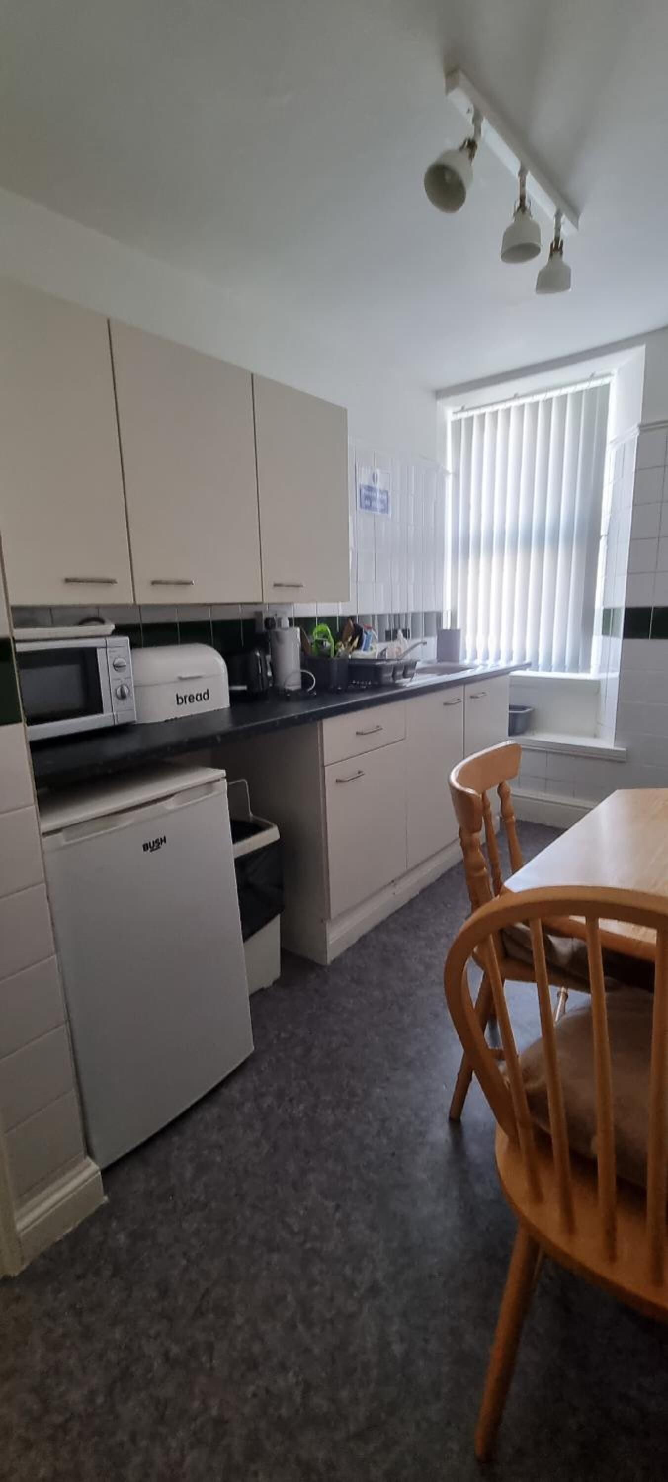 A compact kitchen with white cabinets, a bread maker, a mini fridge, and a dining area with a wooden table and chairs.