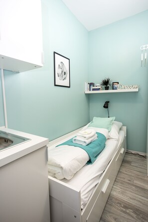 A bedroom with a bed, a bedside table, a wall-mounted shelf with books and a plant, and a framed picture on the wall. - Micro Appartments Katz (Nuremberg)