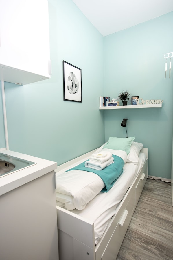 Un dormitorio con cama, una mesita de noche, estante con libros y una planta, y un cuadro en la pared.