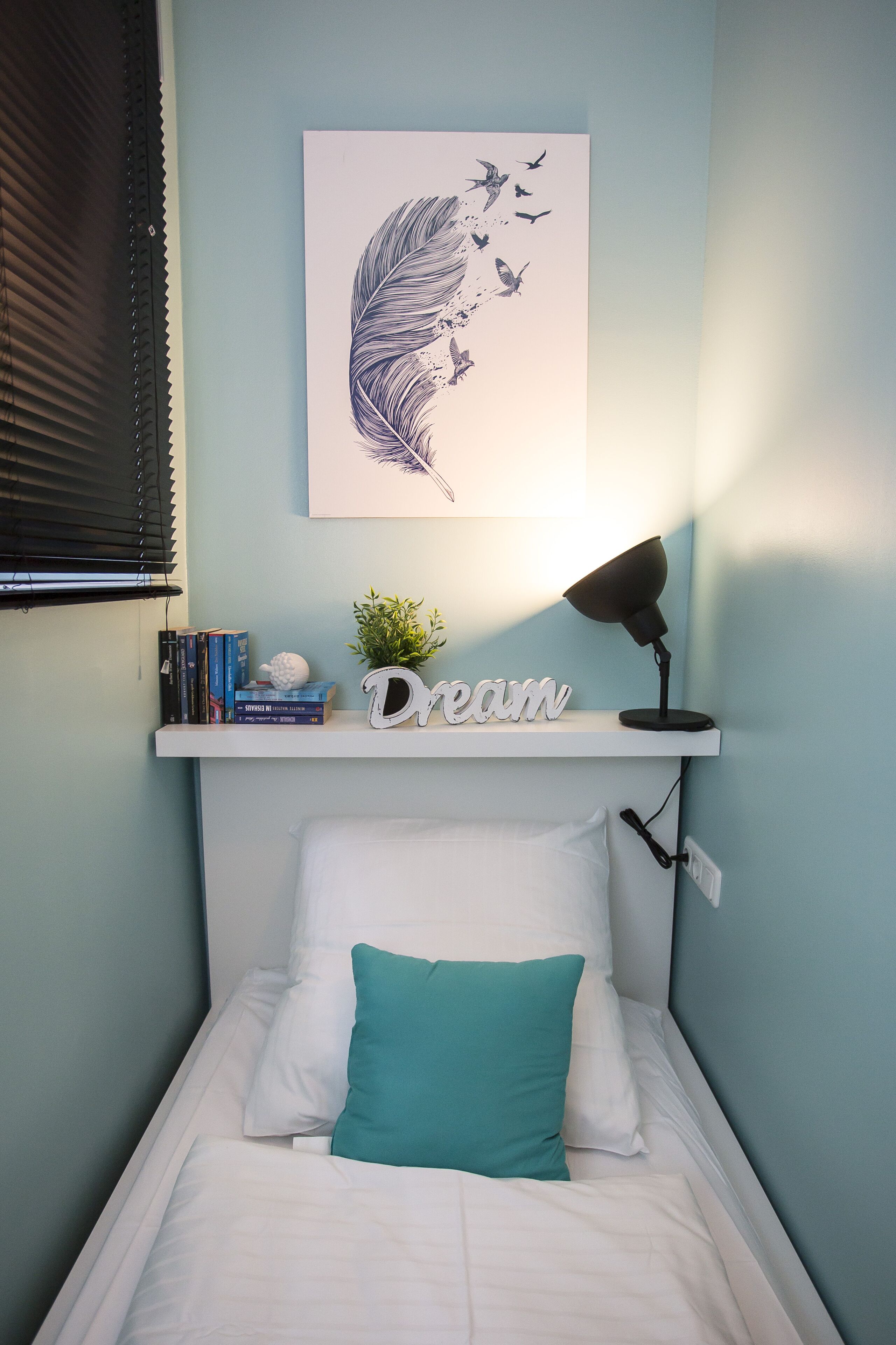 Cama bem arrumada com um travesseiro azul-turquesa e um travesseiro branco, uma pequena estante com livros e uma planta, uma luminária de parede e uma obra de arte emoldurada com pássaros.